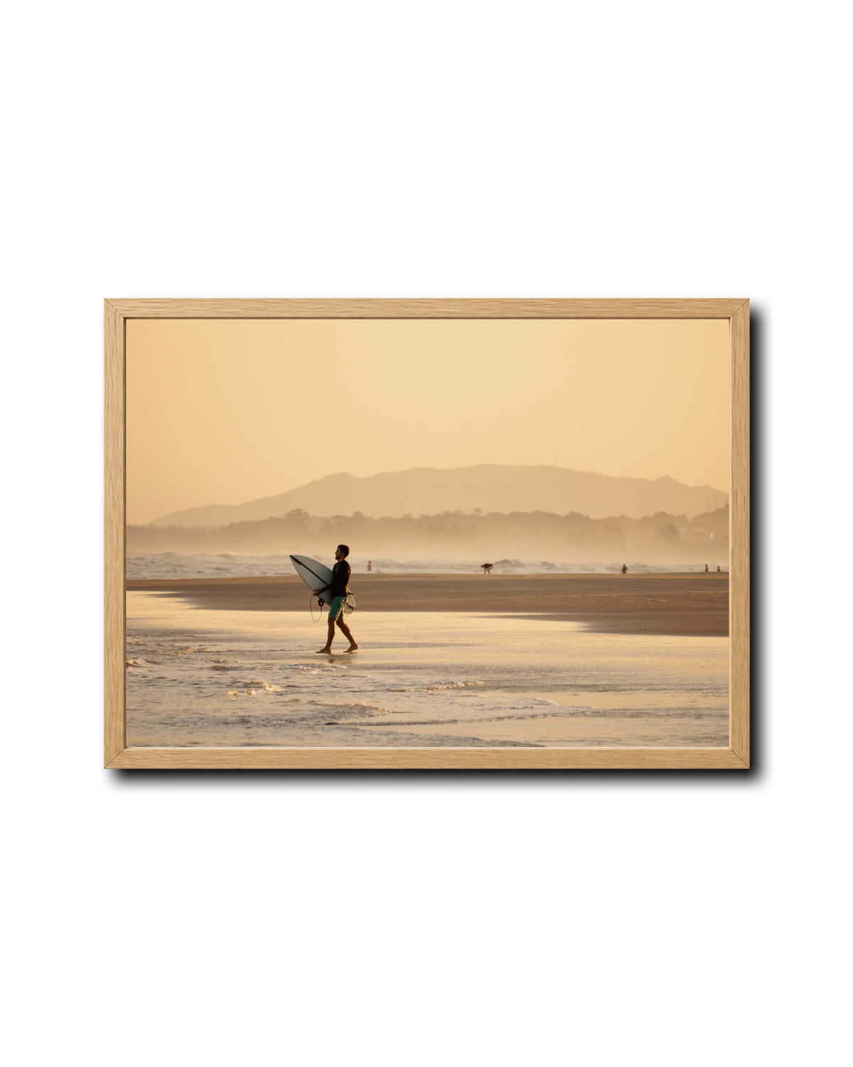 A framed photographic print of a surfer walking on a beach at sunrise, with a warm golden-orange sky and distant mountains. The print is housed in a light natural wood frame and is displayed against a clean white background with a subtle drop shadow.