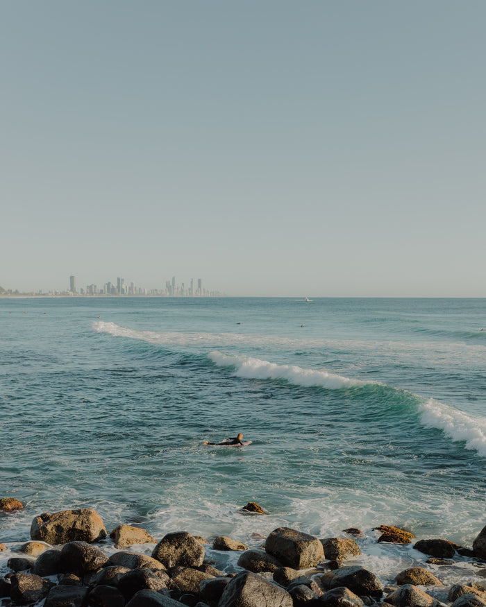 Surfer paddling out at Burleigh with the Gold Coast city skyline behind, photographed by Chris Hepworth