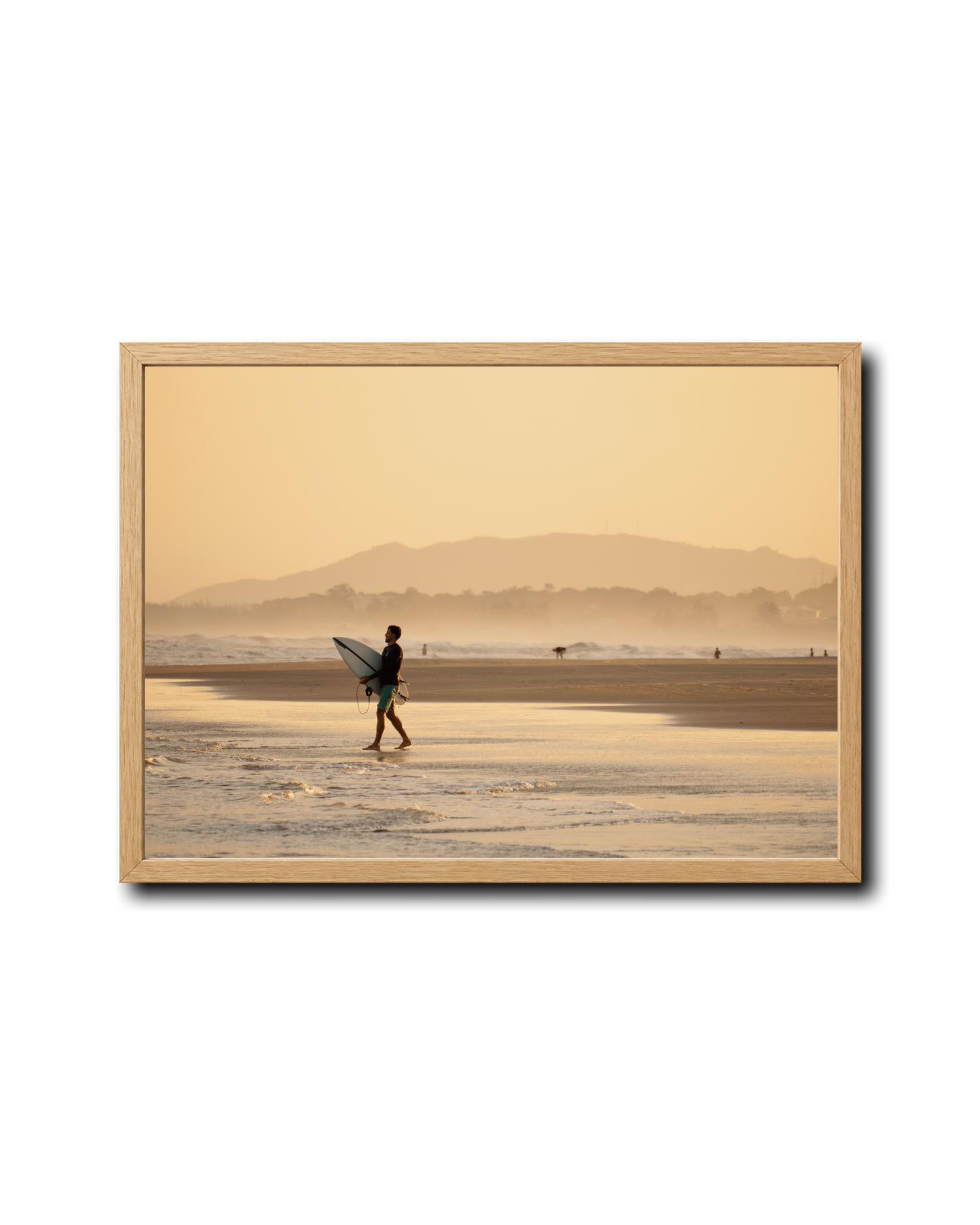 A framed photographic print of a surfer walking on a beach at sunrise, with a warm golden-orange sky and distant mountains. The print is housed in a light natural wood frame and is displayed against a clean white background with a subtle drop shadow.
