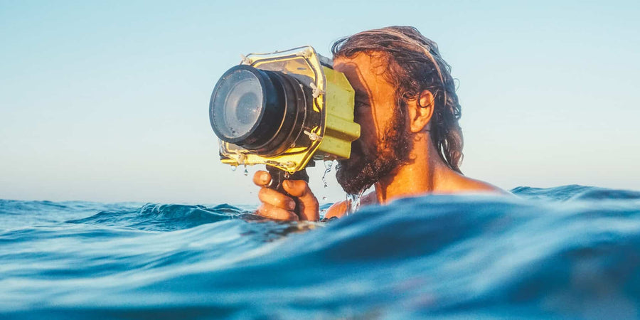 A man in the ocean holding a waterproof camera housing above the water surface, with blue water and sky in the background.