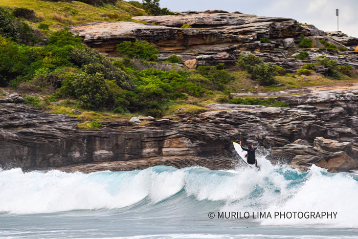 Surfer riding a wave beneath rocky sandstone cliffs, photographed by Murilo Lima