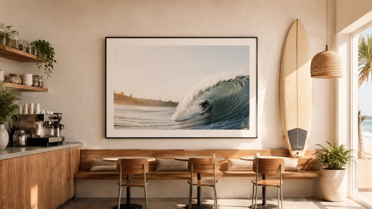 Framed surf photography print displayed in a sunlit coastal café beside a surfboard.